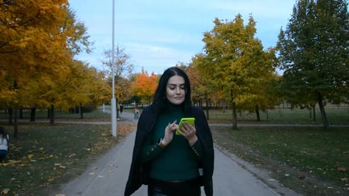 A Beautiful Brunette Girl with a Phone in Her Hands Writes a Message to Friends and Walks Along the