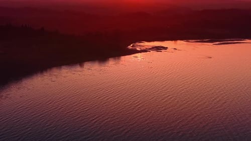 Paisaje acuático estriado de color rosa al atardecer. La costa boscosa apenas se ve a la sombra. Vista aérea.