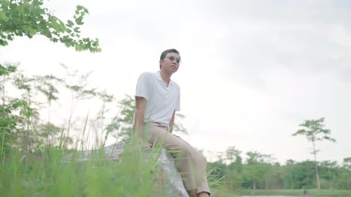 Man rests and relaxes, sitting on a large rock by the water. Look at nature and smile happily