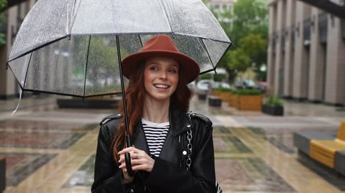Portrait of Happy Young Redhead Woman in Hat Walking on Street in City with Transparent Umbrella