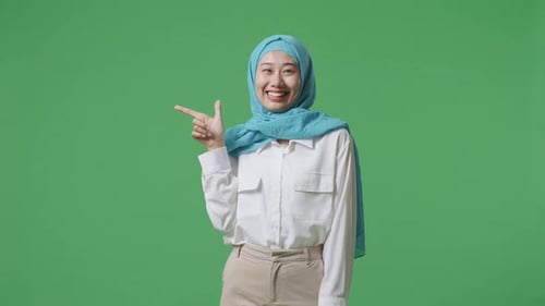 Asian Muslim Woman Smiling And Pointing To Side While Standing In The Green Screen Background