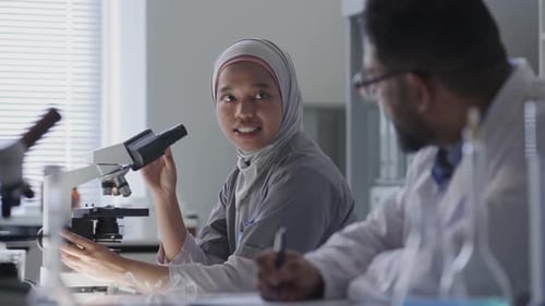 Female Scientist in Hijab Using Microscope and Speaking with Colleague in Lab