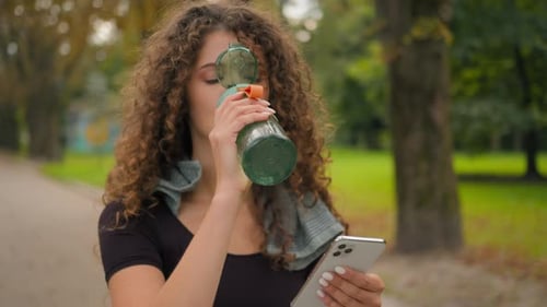 Woman Drinks Water Checking Smartphone in Park
