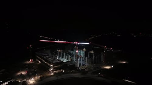 Night aerial drone view of construction site with illuminated cranes and building framework,