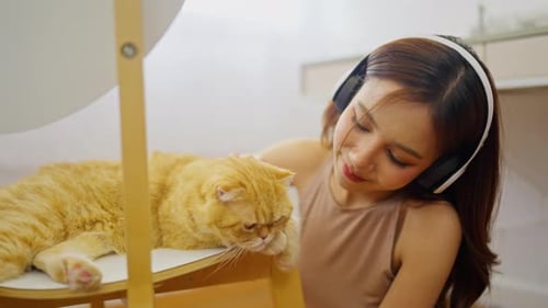 Pretty Asian woman with headphone sit on the floor near orange cat and she smile with happiness
