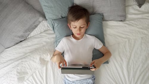 A cheerful young boy is playing with a tablet computer while lying on his bed during the day