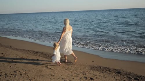 Mom and Little Daughter in White Dresses Walking on the Sea Beach at Sunset