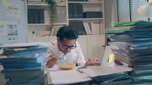 Young Adult Works at Desk Eating Noodles