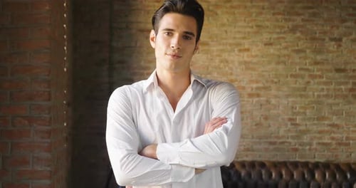A handsome man in his loft dresses his sexy white shirt in a sexy way, looks into his room and smi