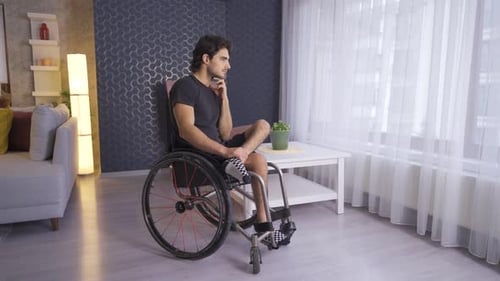 Disabled young man is thoughtful and looks out the window of the house.