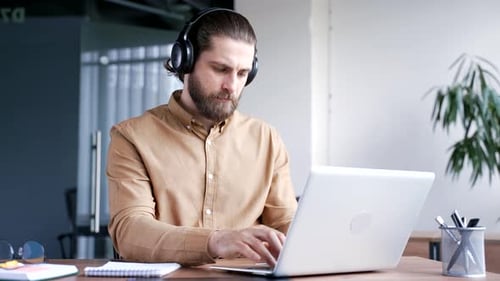 Focused businessman or IT specialist in wireless headphones works on a laptop sitting at a workplace