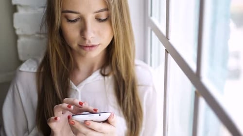Smiling Young Woman Using Phone by Window