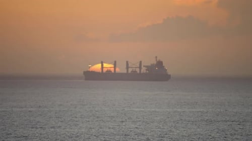 Wide cinematic shot of a cargo ship carrying cargo as it enters a port to offload with the sun setti