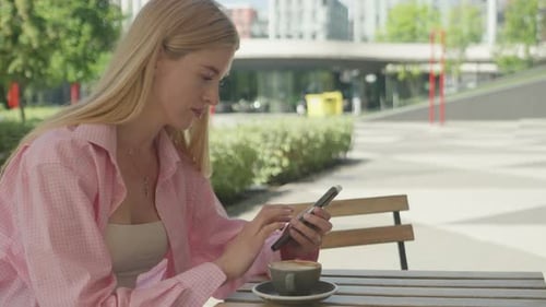 Blonde Woman Texting On Smartphone At Outdoor Cafe Table With Coffee