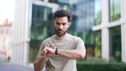 Adult athlete runner looking smart watch standing near a modern urban building on a city street.