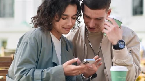 A happy couple outdoors near a cafe. Looking in the phone, smiling, talking, coffee. Autumn atmosphe