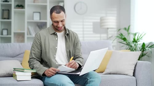 Man Studies at Home with Laptop and Notebook