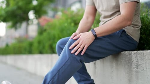 Male hands massaging knee leg muscles sitting on city park alley. Man athlete suffers from painful