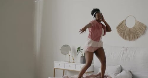 Slow Motion Portrait of Cheerful Girl Wearing Headphones Jumping and Dancing on Bed in Apartment