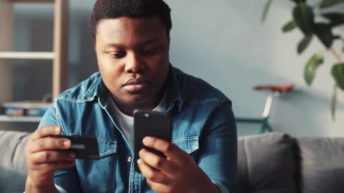 Close Up Serious Young African American Man Use Phone and Credit Card Sit on Sofa in Home Online