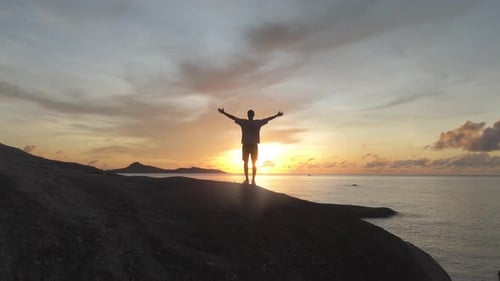 Man Stands at Ocean Shore Orange Sunset Raising Arms in Enjoyment of Moment Majestic Nature