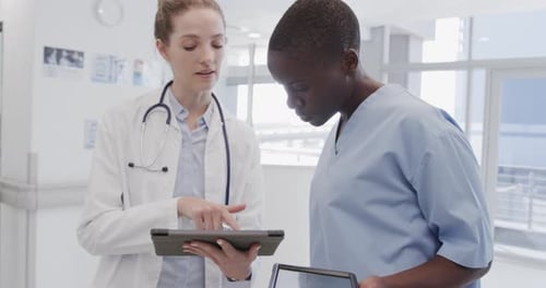 Diverse female doctor and nurse looking at tablets and talking in hospital corridor, in slow motion