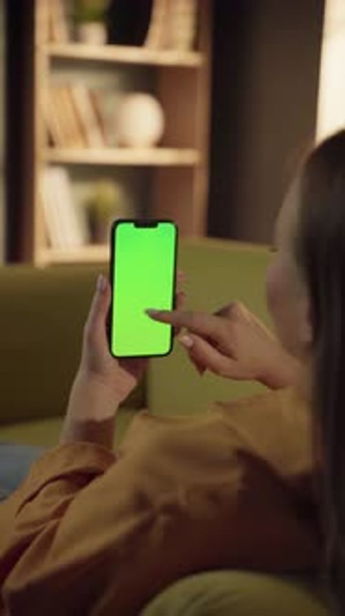 Woman on a Couch Holding a Vertical Smartphone with a Green Screen Tapping and Scrolling with Her