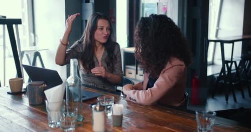 Dos jóvenes hermosas amigas multiétnicas, mujeres de negocios, hablan casualmente usando una computadora portátil trabajando juntas