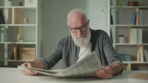 Ein ernster alter Mann mit Brille sitzt an einem Tisch und liest eine Zeitung