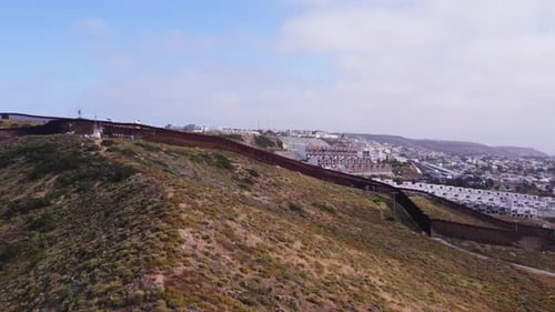 Drone Flight Over Hillside U.S.–Mexico Border Fence with Urban Background