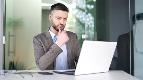 Serious thoughtful businessman works on a laptop sitting at a workplace in a business office.