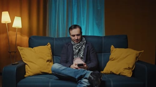 Young Caucasian Man Relaxing on Couch Using Smart Phonetyping Message in Modern House