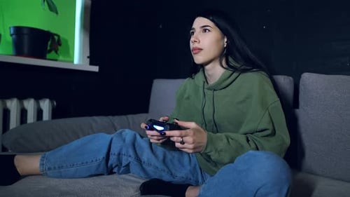 Eine junge erwachsene Frau sitzt nachts zu Hause auf einem Sofa und spielt ein Videospiel auf der Konsole