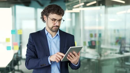Confident serious businessman is using digital tablet while standing in business office. Man worker