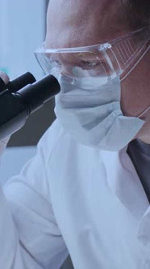Male Lab Technician in Mask and Gloves Examining Samples with Microscope