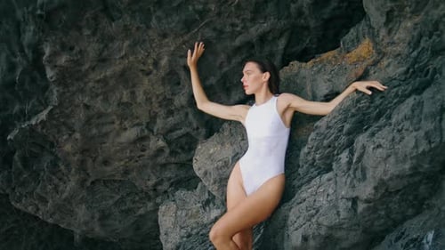 Hot Young Woman Posing in White Elegant Swimsuit Leaning at Beach Rock