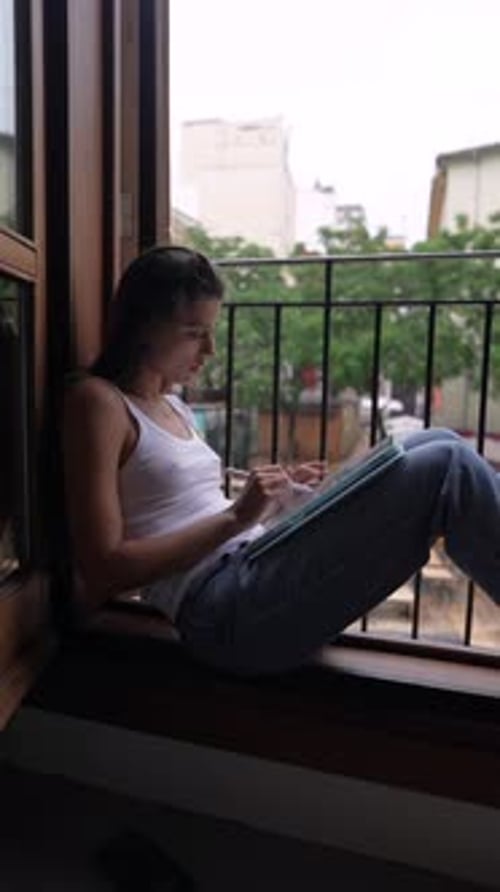 A Young Woman is Peacefully Reading a Tablet By a Window in a Casual Home Setting