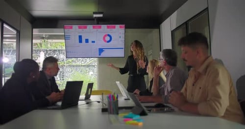 Young presenter pointing at data chart while team listens during meeting in modern office