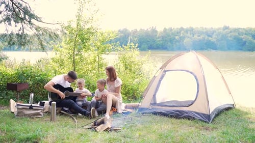camping by lake in woods. happy family dad mom and little kids sitting by fire and tent in nature.