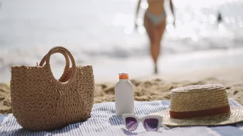 Beach Essentials Laid on Towel Sunscreen Bottle UV Protection Person Walks Towards Sea Prepares for