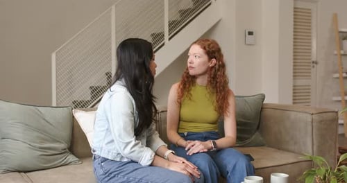 Diverse female friends sitting on sofa at home redhead recounting for help brunette placing hand