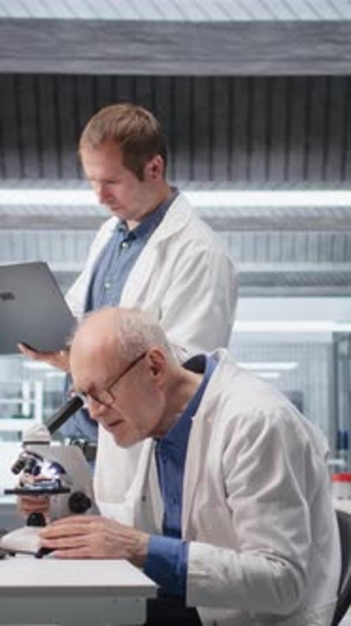Vídeo vertical: Cientista idoso usando microscópio com um colega para estudo clínico