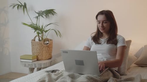 Smiling Brunette arbeitet an ihrem Laptop, während sie im Bett liegt. Ein junges Mädchen arbeitet von zu Hause aus