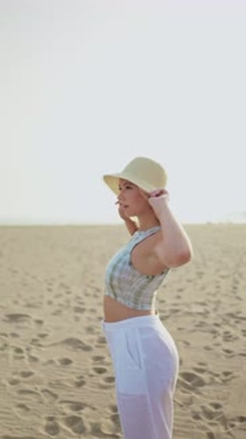 Vertical video of a young woman putting her hat on while enjoying her holiday on the beach