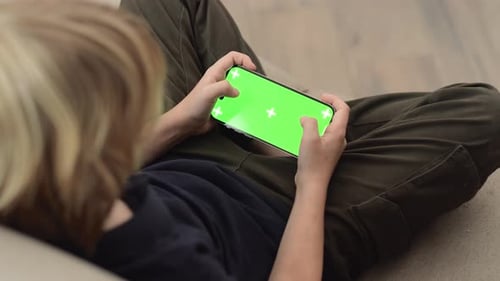 Chroma-Key-Handy mit grünem Bildschirm in Kinderhänden, nicht wiederzuerkennen, blonder Junge spielt Telefon in der Hand