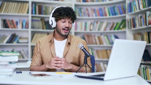 Young happy university teacher in wireless headphones is recording a podcast using a laptop