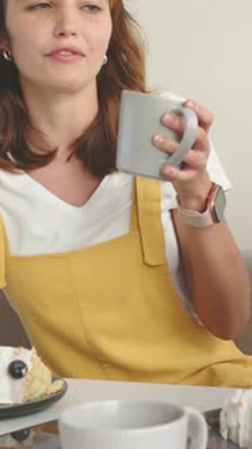 Vertical video: Holding gray mug, woman in yellow overalls accepting fork offered with cake at home