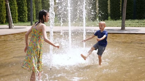 Two Cheerful Children Play in the Fountain in the Summer in the City Park