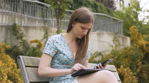 A Beautiful Young Caucasian Woman Works on a Tablet and a Smartphone As She Sits on a Bench