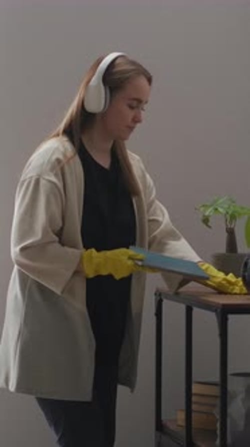 Young Woman Wearing Headphones and Rubber Gloves Cleaning a Small Indoor Plant Shelf Carefully